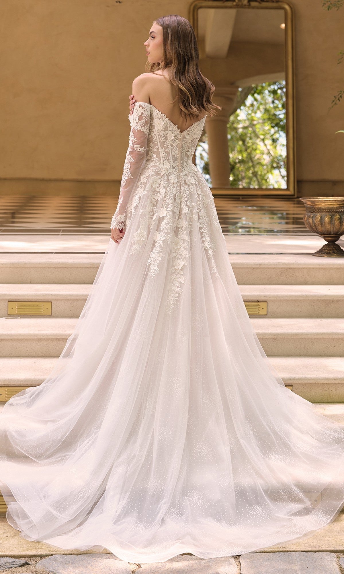 A woman stands on steps, facing away, wearing the Andrea and Leo WL067 White Lace Wedding Dress with off-the-shoulder 3D floral lace applique and a corset bodice. A large mirror and lush greenery fill the background.